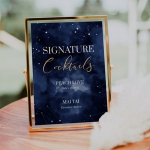 May include: A gold-framed sign with a navy blue background and white stars. The sign reads "Signature Cocktails" in gold lettering. Below the title, it lists "Peach Love" as the bride's choice and "Mai Tai" as the groom's choice.