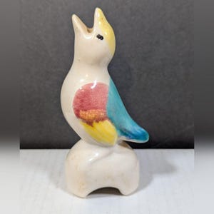 May include: A ceramic bird figurine with a cream-colored body, yellow head, and blue wing. The bird has a red and orange patch on its chest and is perched on a white base. The bird is in a singing pose.
