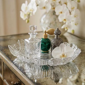 May include: A clear glass decorative tray holds a crystal perfume bottle, a green malachite perfume bottle, and a silver perfume bottle. A white orchid flower rests on the tray. A vase of white orchids is in the background.