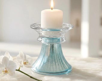 May include: A clear glass candle holder with a flared rim and a textured, light blue base. A white pillar candle sits inside the holder with a lit flame. A white orchid flower with a green stem is positioned to the left of the candle holder.