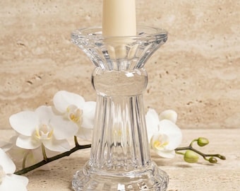 May include: A clear glass candle holder with a fluted, vertical ribbed design and a wide, scalloped base. The holder features a thick, transparent stem and a flared top rim, currently holding a single, solid cream-colored taper candle.