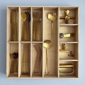 May include: A wooden drawer organiser with compartments for storing gold-toned cutlery, including spoons, forks, knives, and straws. The organiser has a light wood finish and is divided into multiple sections.