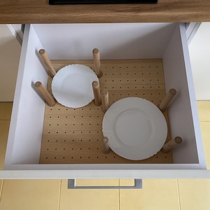 May include: A white kitchen drawer with a wooden insert and a silver handle. The insert has a grid of holes and two white plates are stacked on top of wooden dowels.