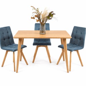 May include: A rectangular light wood dining table with four tapered legs and three blue upholstered chairs featuring tufted backrests. A clear glass vase containing dried botanical stems sits in the center of the table surface.