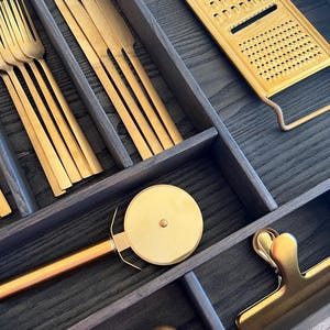 May include: A black drawer organiser with gold utensils, a gold grater, a gold pizza cutter, and gold measuring spoons.