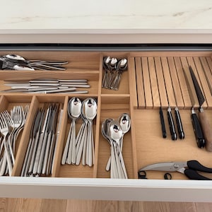 Peut inclure: Un organisateur de tiroir de cuisine en bois avec plusieurs compartiments contenant des fourchettes, des cuillères et des couteaux en acier inoxydable. Il comprend une section dédiée avec des fentes verticales pour les couteaux et des ciseaux de cuisine.
