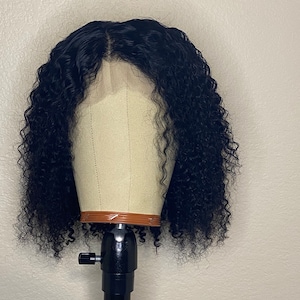 Peut inclure: Perruque noire bouclée présentée sur une tête de mannequin. La perruque présente une ligne de cheveux naturelle et une raie profonde. Les cheveux sont densément bouclés, créant un aspect volumineux. La tête de mannequin est beige clair, montée sur un support noir réglable.