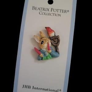 May include: A colorful brooch featuring a rabbit character holding carrots, with a red bird perched on a branch. The brooch is on a card with the text "BEATRIX POTTER COLLECTION" and "JHB International".