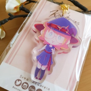 Peut inclure: Un porte-clés en acrylique transparent représentant un personnage de dessin animé avec un chapeau et une robe violets, portant des lunettes. Le porte-clés est attaché à un fermoir doré. L'emballage comprend le texte "Srta. Acuarela" et des icônes de médias sociaux.