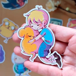 May include: A die-cut vinyl sticker featuring a cartoon illustration of a blonde character with pink-tinted hair wearing a blue shirt and purple shorts, embracing a round, orange, and pink creature with closed eyes in a warm, affectionate pose.