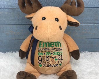 May include: A tan plush moose toy with dark brown antlers and hooves, featuring a navy blue ribbon around its neck. The chest is embroidered with the name Emeth, Surafel Argaw, the date 06-25-2022, 9 pounds 5 ounces, 21 inches, and 7:56 AM.