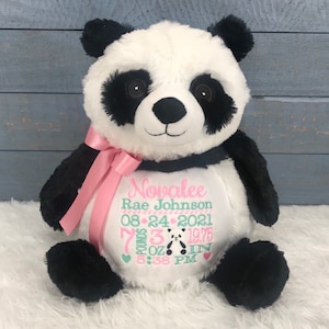 Może przedstawiać: Pluszowa zabawka panda z czarno-białym futrem, z r&oacute;żową satynową wstążką zawiązaną wok&oacute;ł szyi. Biały brzuch jest wyhaftowany imieniem Novalee Rae Johnson, datą 24-08-2021 oraz szczeg&oacute;łami urodzenia, w tym 3,26 kg i 50,17 cm.