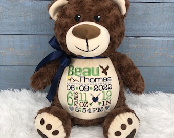 May include: A brown plush teddy bear with a cream-colored belly featuring embroidered text that reads Beau Thomas, 06 09 2022, 6 pounds 11 oz, 19 in, and 5:54 PM, accented with a dark blue ribbon tied around its neck and dark brown paw pads.