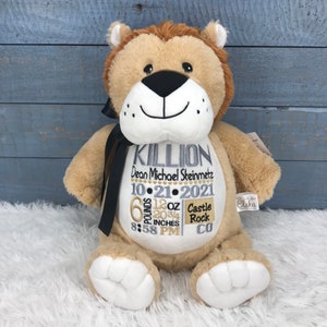 May include: A plush lion toy with embroidered details. The lion is tan with a brown mane and a black bow. The front of the lion has embroidered text with a name, date, time, weight, and location. The lion is sitting on a white surface.