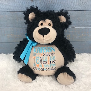 May include: Black and tan plush bear with blue ribbon. Embroidered on the bear's chest is the name "Karson Xavier", birth stats, and the date "07.29.2022".