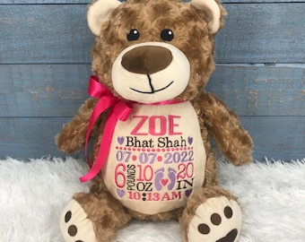 May include: A brown plush teddy bear with a cream-colored belly featuring embroidered text ZOE, Bhat Shah, 07-07-2022, 6 pounds 10 oz, 20 IN, and 10:13 AM, accented with purple hearts, pink footprints, and a bright pink satin ribbon around its neck.