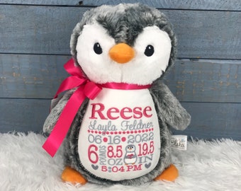 May include: A plush penguin toy with grey and white fur, an orange beak, and orange feet. A bright pink ribbon is tied around its neck. The white belly features embroidered text: Reese, Layla Feldner, 06-16-2022, 6 pounds 8.5 oz, 19.5 in, 5:04 PM.