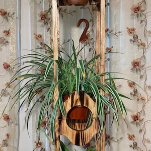 Op de afbeelding: Een houten plantenstandaard met drie niveaus, elk met een potplant. De standaard heeft een rustieke, verbrande houten afwerking. De planten zijn weelderig en groen, met lange, hangende bladeren. Een decoratieve metalen haak hangt in het midden.