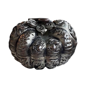 Puede incluir: Un objeto decorativo plateado y ornamentado con forma de calabaza. La superficie está cubierta de patrones y remolinos detallados, con un pequeño elemento cilíndrico de color marrón oscuro en la parte superior. El objeto está sobre un fondo blanco liso.