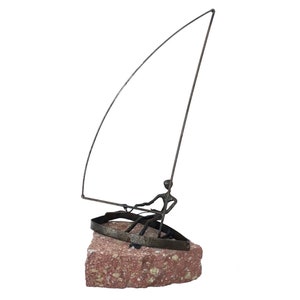May include: A metal sculpture of a sailboat with a figure at the helm, mounted on a reddish-brown stone base. The sailboat has a curved mast and a simple boat design. The sculpture is made of dark metal.