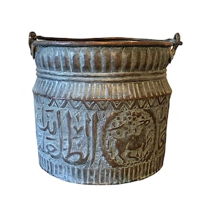 May include: An antique copper bucket with intricate engraved Arabic script and a stylized animal design. The bucket has two handles and a worn, aged patina.