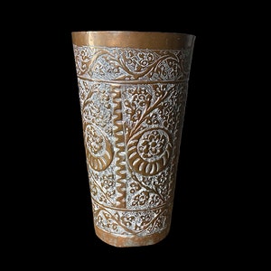 May include: A tall, brass-colored metal cup with intricate floral and scrollwork patterns. The cup has a slightly flared shape and a detailed, textured surface. The design features repeating motifs of flowers and vines, creating a decorative and ornate appearance.