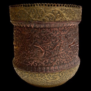Puede incluir: Un macetero grande, redondo, de cobre y oro con intrincados diseños florales y animales. El macetero tiene una base ancha y redonda y una parte superior estrecha.