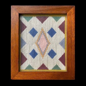 May include: A framed needlepoint piece with a geometric design featuring a pink diamond in the center surrounded by blue and green diamonds. The frame is made of dark wood.