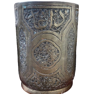May include: A vintage, octagonal, brass cup with intricate engraved designs. The cup features a central circular design with a knotwork pattern and Arabic script around the rim.
