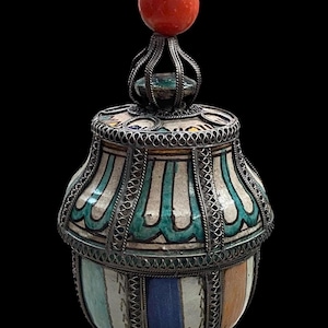 May include: A decorative ceramic jar with a silver filigree frame and a red, spherical top. The jar features a colorful design with blue, white, and orange vertical stripes. The lid is adorned with a silver filigree crown.