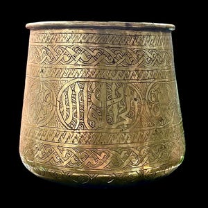 Peut inclure: Un récipient antique de couleur laiton, orné de motifs gravés complexes. Le récipient présente des bandes de motifs géométriques et calligraphiques, avec une bande centrale plus large contenant une écriture ornée. La forme est légèrement conique.