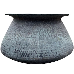 May include: A weathered, dark gray metal pot with a textured, scale-like pattern. The pot has a wide, rounded body and a flared rim. The pot's surface shows signs of age and wear, suggesting it is an antique or vintage item.