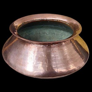 May include: A copper pot with a hammered finish, featuring a rounded base and a flared rim. The interior has a green tint, contrasting with the polished copper. The pot is set against a black backdrop.