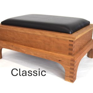 May include: A wooden storage ottoman with a black leather top. The ottoman has a classic design with a simple rectangular shape and four legs. The wood is a light brown color and the legs are slightly curved.