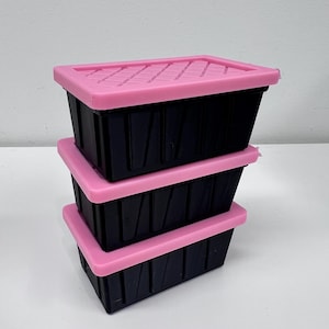 May include: Three black rectangular storage containers stacked on top of each other, each with a pink lid. The lids have a ridged design. The containers are on a white surface.