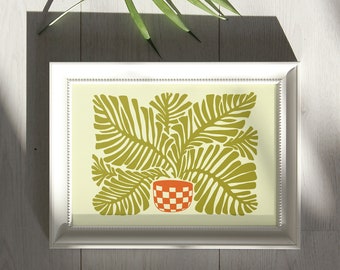 May include: A framed art print featuring a stylized illustration of a potted plant with large, green leaves. The pot is orange and white checkered. The print is in a white frame, resting on a light wood surface. A green leaf casts a shadow.