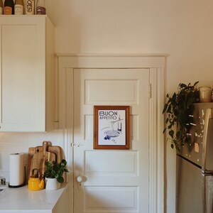 Puede incluir: Una cocina blanca con una puerta blanca con una impresi&oacute;n enmarcada que dice "BUON APPETITO" en letras azules. Hay una tetera amarilla en el mostrador y una planta en una maceta blanca.