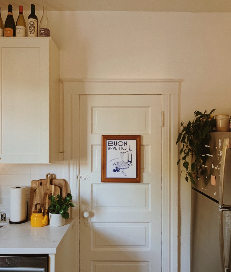 Puede incluir: Una cocina blanca con una puerta blanca con una impresi&oacute;n enmarcada que dice "BUON APPETITO" en letras azules. Hay una tetera amarilla en el mostrador y una planta en una maceta blanca.