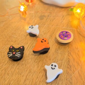 May include: Five Halloween-themed charms, including a black cat, a white ghost, an orange ghost, a donut with purple frosting and sprinkles, and another white ghost. The charms are on a wooden surface.