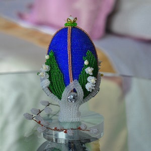 May include: A blue and green beaded Easter egg with white flowers and a crystal top, sitting on a clear stand.