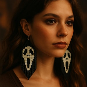 Puede incluir: Pendientes de cuentas en blanco y negro con diseño de máscara de Scream. Los pendientes tienen forma rectangular y larga, con flecos en la parte inferior. La máscara es blanca y el fondo negro. Cuelgan de ganchos dorados.