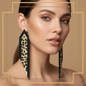 Pode incluir: Close-up de uma pessoa usando brincos longos de contas. Os brincos são pretos, brancos e bege, com um padrão geométrico. São triangulares no topo e têm franjas longas. O fundo é bege neutro.