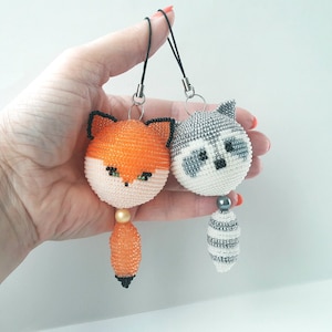 May include: Two beaded animal charms, a fox and a raccoon, with black eyes and white accents. The charms are attached to black cords with silver rings.