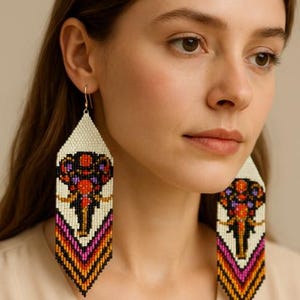 May include: Long, beaded earrings featuring an elephant design. The earrings have a cream-coloured background with a colourful elephant motif in red, purple, orange, and black. The lower section has a geometric pattern in pink, orange, and black.