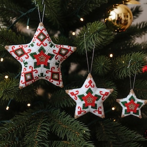 May include: Three beaded star decorations hang from a Christmas tree. The stars are white with red, green, and gold beadwork in a geometric pattern. A gold bauble is visible in the background.