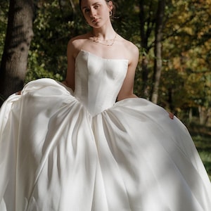 May include: A woman wearing a white strapless wedding dress with a sweetheart neckline. The dress has a full skirt and is made of a smooth, satin fabric. The woman is standing in a forest setting with trees and leaves in the background.