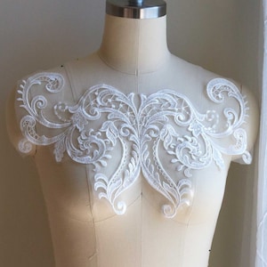 May include: White lace applique with a delicate floral design. The applique is displayed on a dress form.