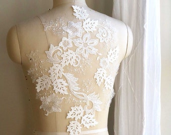 May include: A white floral lace applique is pinned to a mannequin. The applique features a detailed design of flowers and leaves. The delicate lace is semi-transparent, allowing the mannequin's form to be partially visible. The design is symmetrical and elegant.