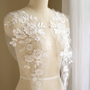 May include: White lace applique with floral designs and bead embellishments. The applique is displayed on a dress form.
