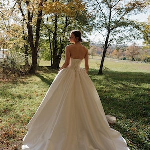 May include: A white strapless wedding gown with a fitted bodice and a full skirt. The gown has a lace-up back and is made of a smooth, satin fabric.